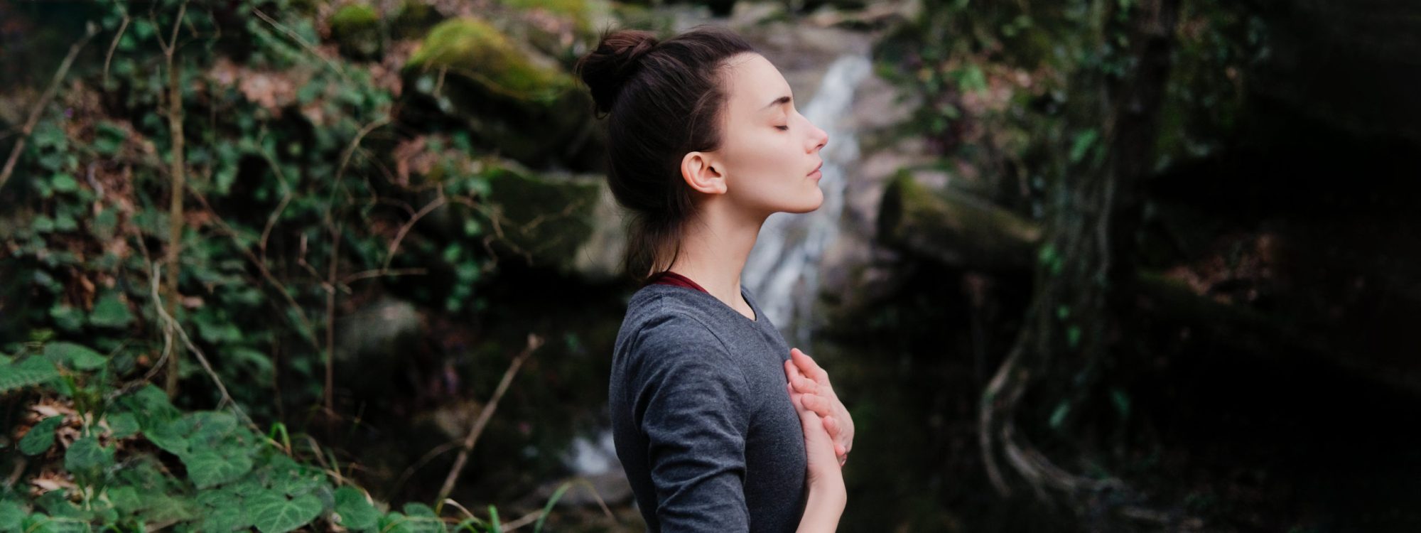 Young woman practicing breathing yoga pranayama outdoors in moss forest on background of waterfall. Unity with nature concept.